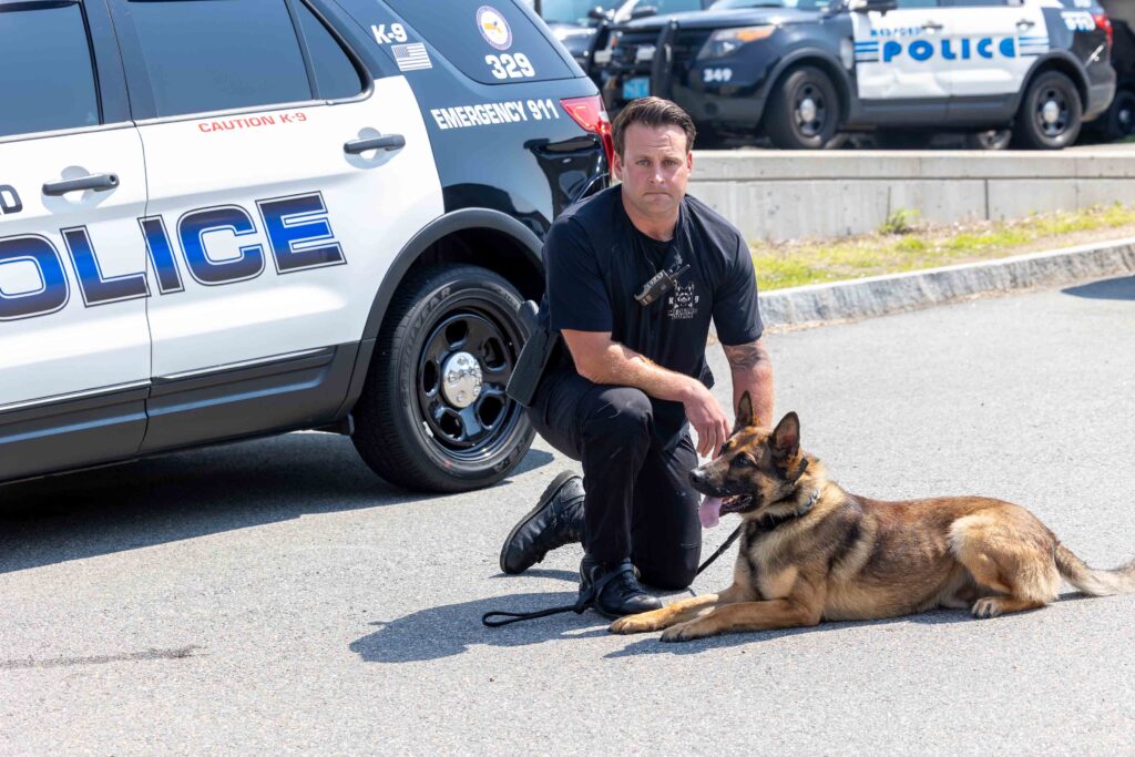 A police officer kneels with a K9 next to a cruiser.